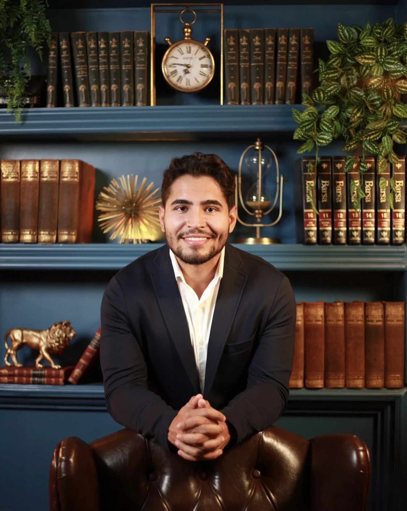 Homem em retrato profissional vestindo terno escuro e camisa branca, sentado em poltrona de couro, com estante de livros e objetos decorativos ao fundo em ambiente sofisticado.