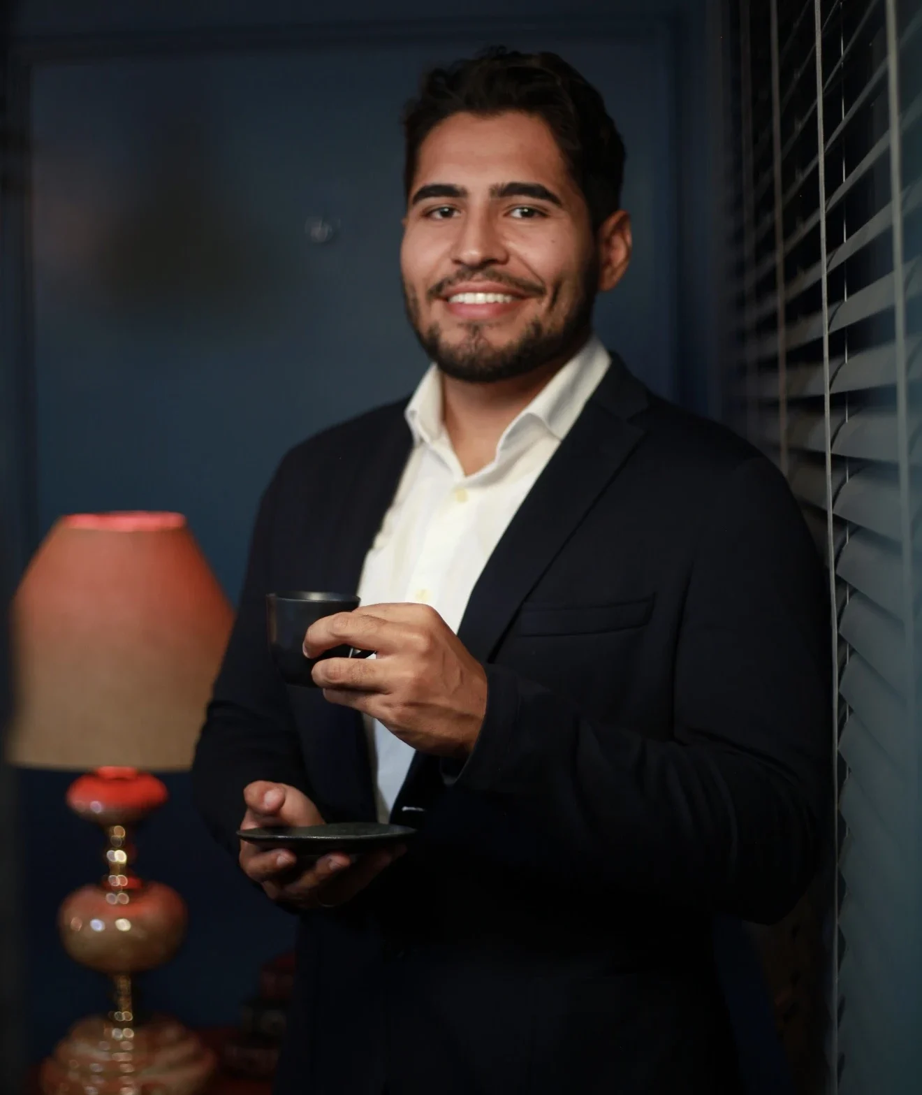 Homem em retrato profissional usando terno escuro e camisa branca, sorrindo enquanto segura uma xícara de café em ambiente corporativo elegante.